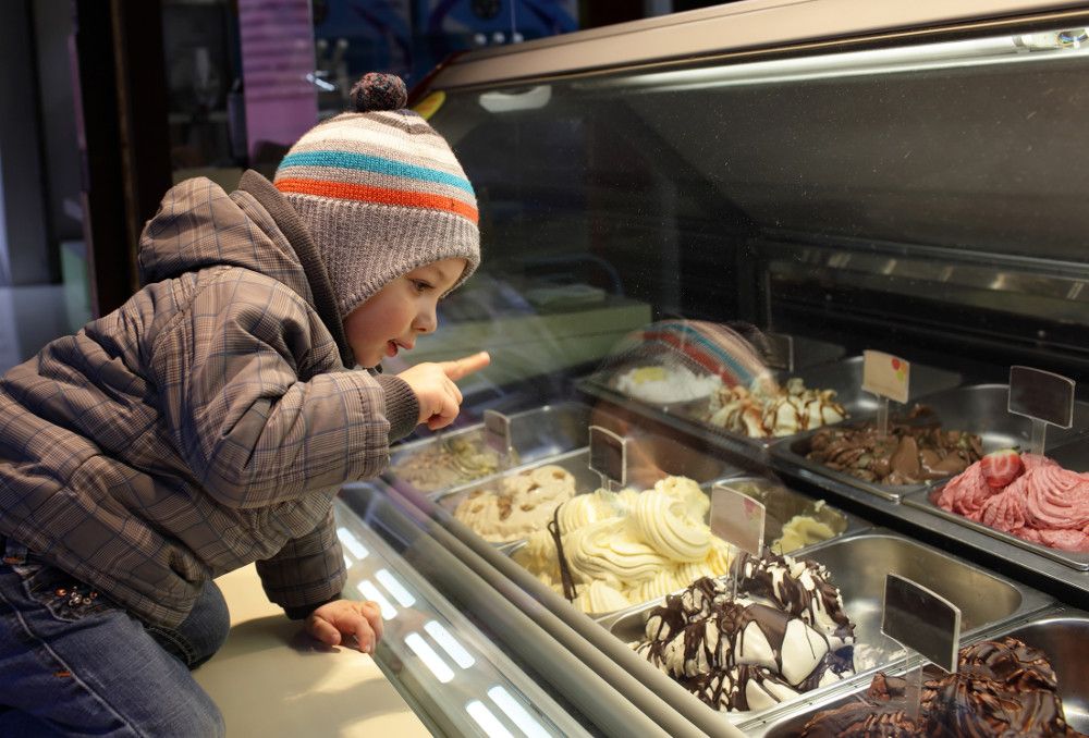 A child in winter wear points at ice cream flavors in a freezer case.